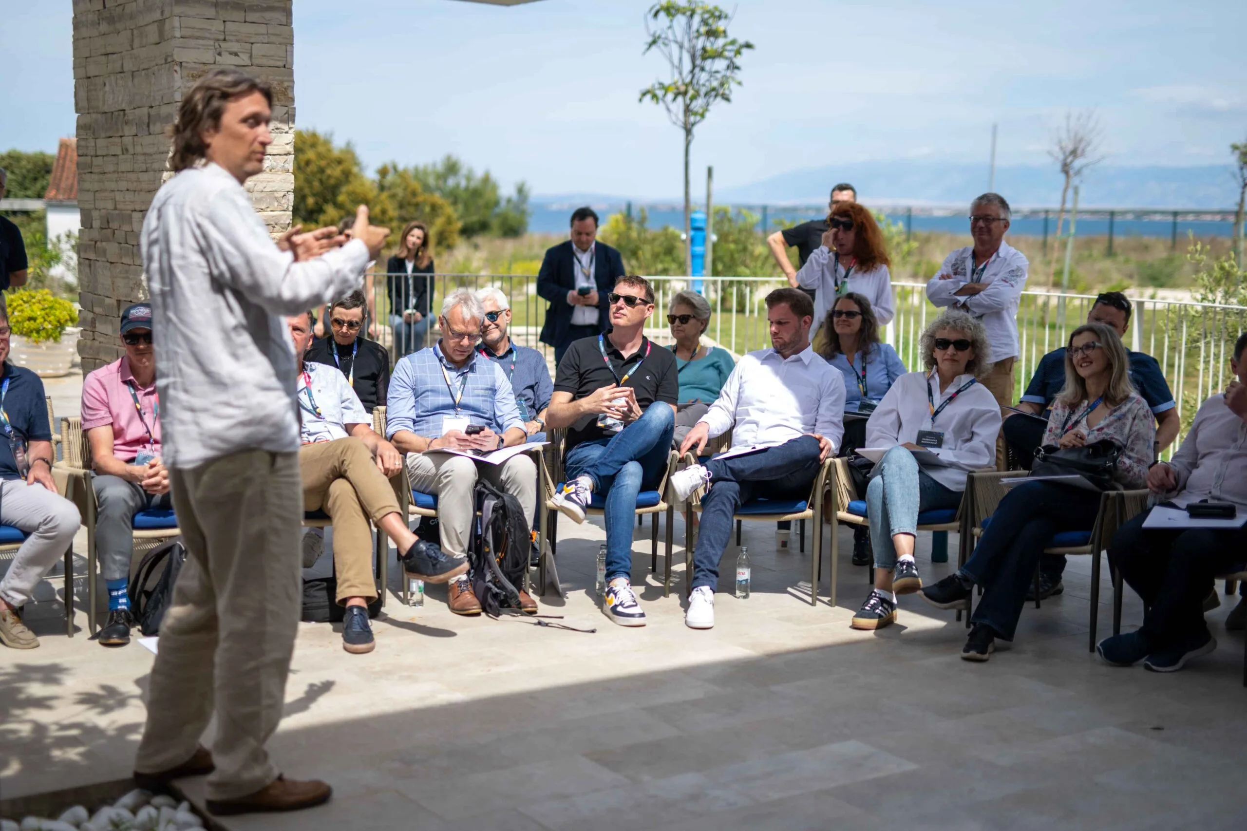 A speaker leads an outdoor session for a seated audience, with the sea behind them